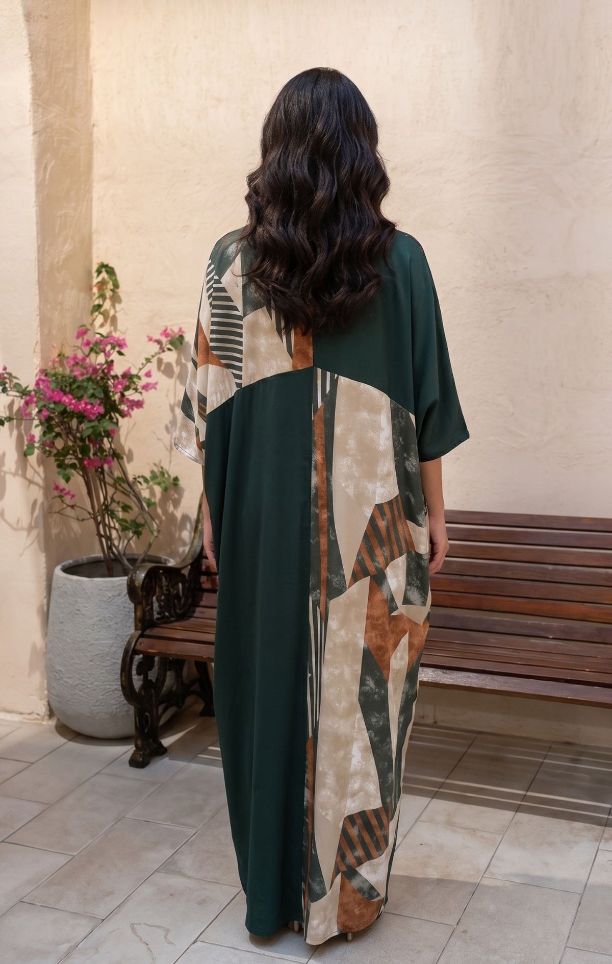 A woman viewed from behind wearing a long, loose kaftan dress with a dark green base and a large abstract geometric panel in beige, brown, and gray. She is standing in an outdoor patio setting with a bench and potted plants.