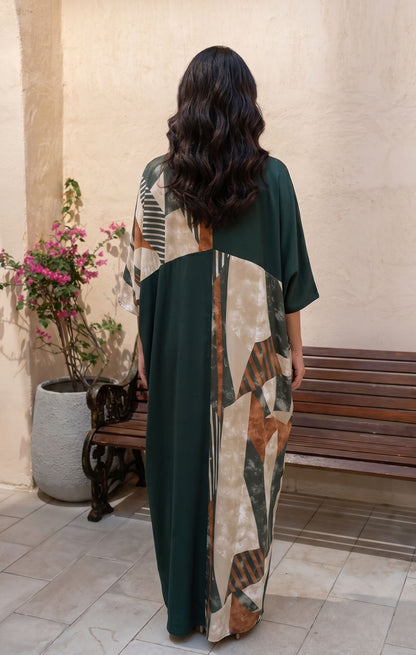 A woman viewed from behind wearing a long, loose kaftan dress with a dark green base and a large abstract geometric panel in beige, brown, and gray. She is standing in an outdoor patio setting with a bench and potted plants.
