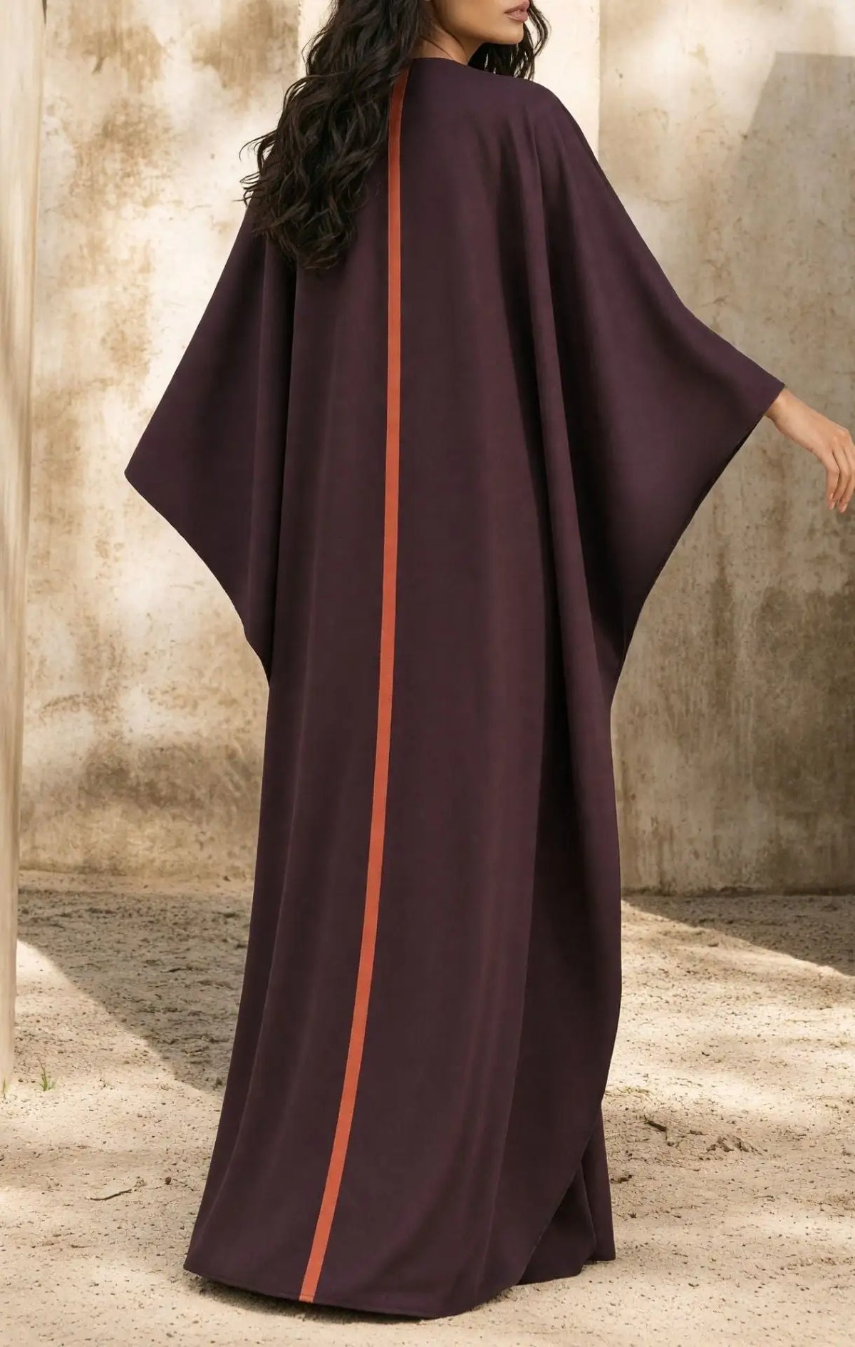 A woman wearing a flowing dark plum kaftan dress with a vertical rust-orange stripe down the center, standing against a textured beige wall.