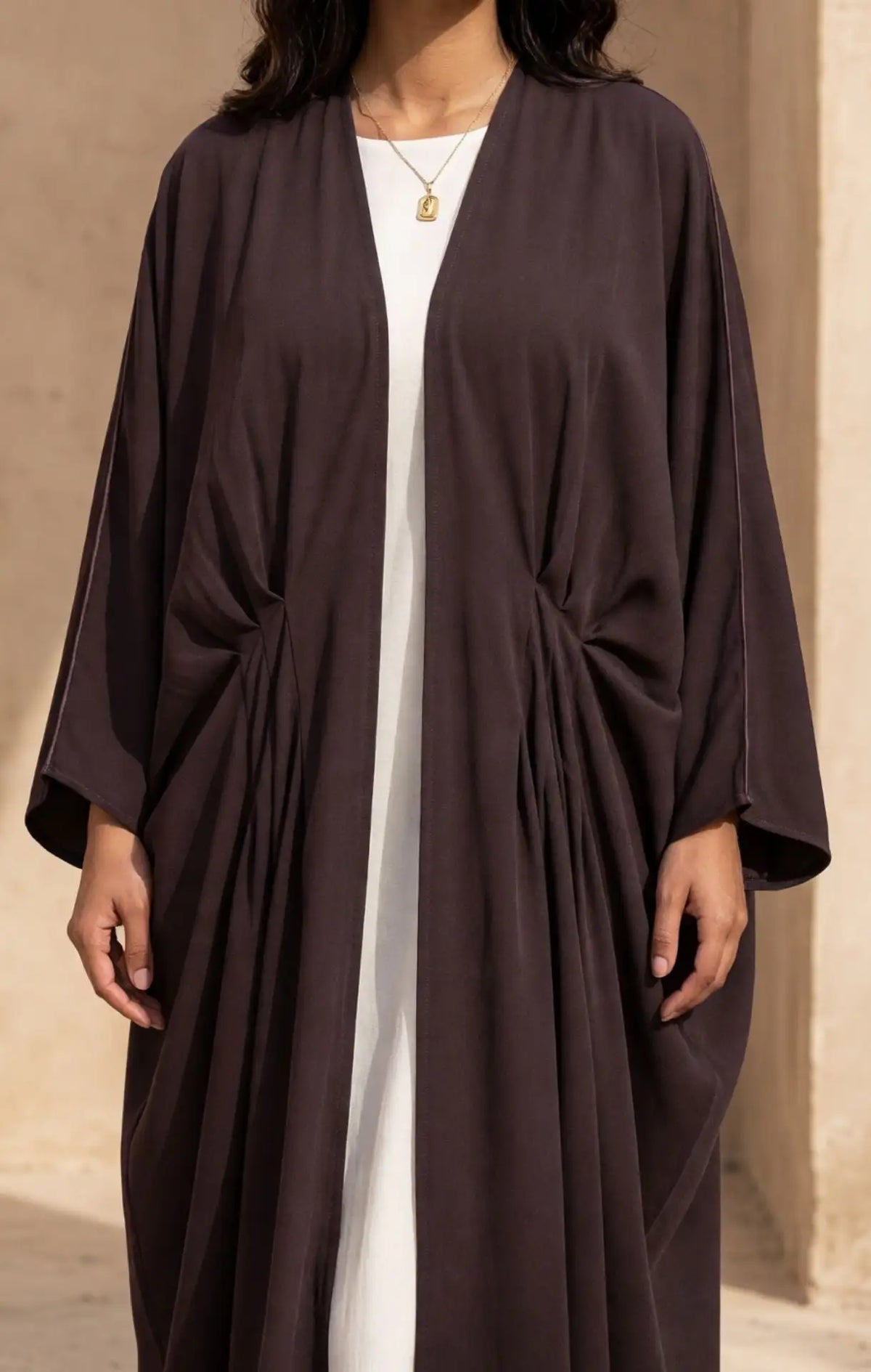 A woman wearing a dark brown, open-front kaftan-style robe with gathered side details over a white dress, set against a warm beige stone background.