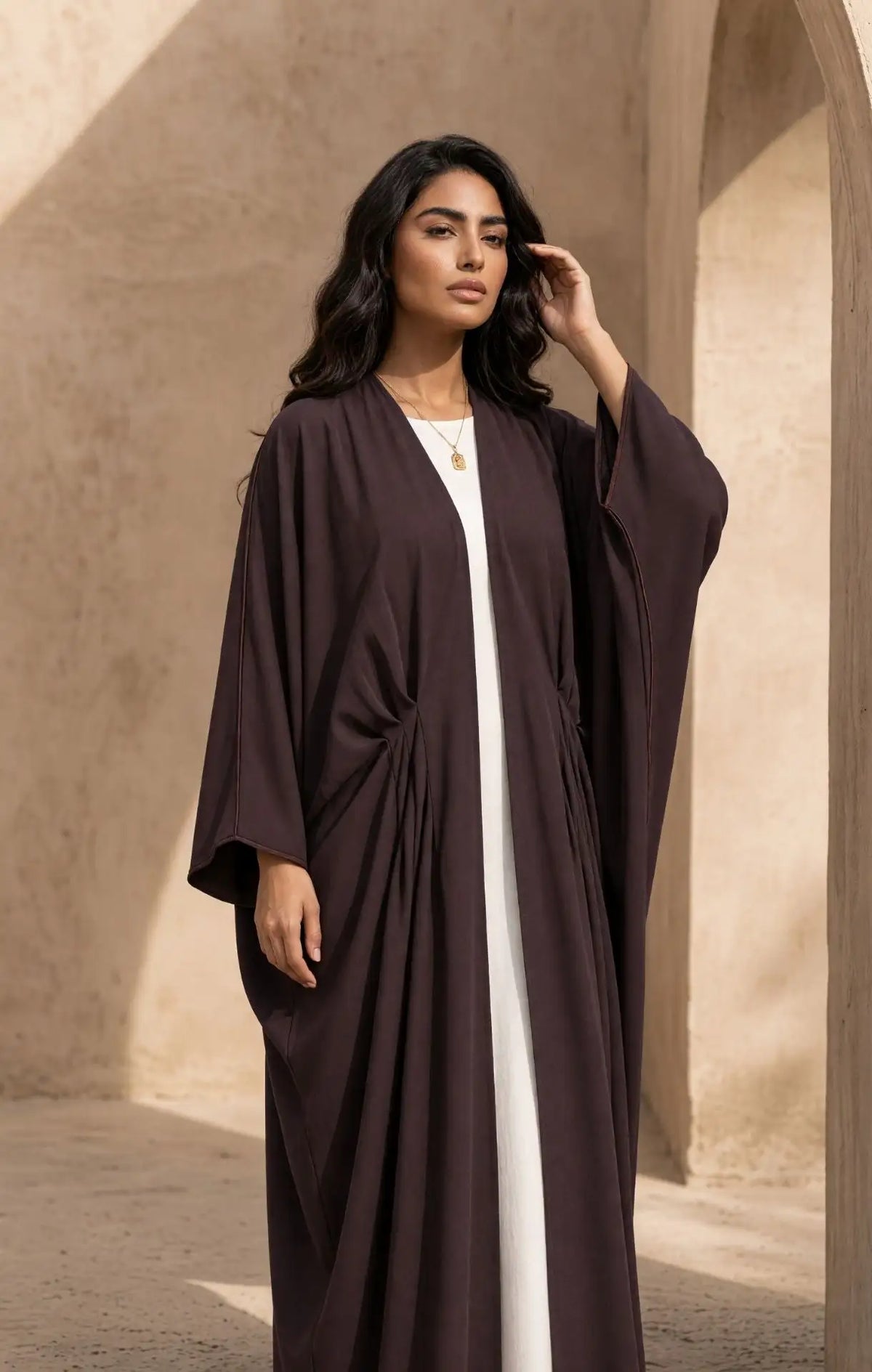 A woman wearing a dark plum abaya with a draped, flowing design over a white inner dress, standing in a warm, sunlit architectural setting with beige walls.