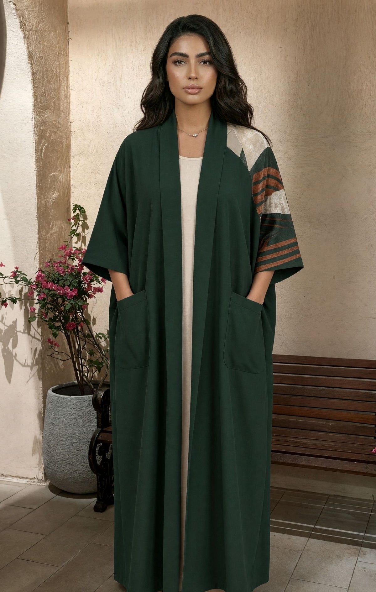 A woman modeling a long dark green open-front abaya with wide sleeves, front patch pockets, and contrasting patterned sleeves in beige, brown, and green, standing in an indoor setting with a bench and potted plant in the background.