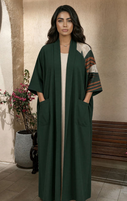 A woman modeling a long dark green open-front abaya with wide sleeves, front patch pockets, and contrasting patterned sleeves in beige, brown, and green, standing in an indoor setting with a bench and potted plant in the background.