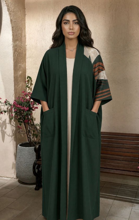 A woman modeling a long dark green open-front abaya with wide sleeves, front patch pockets, and contrasting patterned sleeves in beige, brown, and green, standing in an indoor setting with a bench and potted plant in the background.