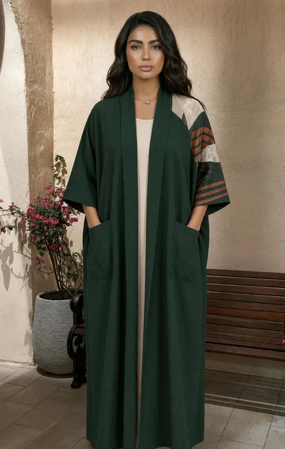 A woman modeling a long dark green open-front abaya with wide sleeves, front patch pockets, and contrasting patterned sleeves in beige, brown, and green, standing in an indoor setting with a bench and potted plant in the background.