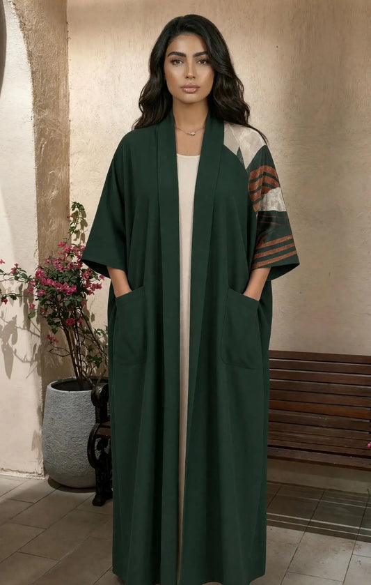 A woman modeling a long dark green open-front abaya with wide sleeves, front patch pockets, and contrasting patterned sleeves in beige, brown, and green, standing in an indoor setting with a bench and potted plant in the background.