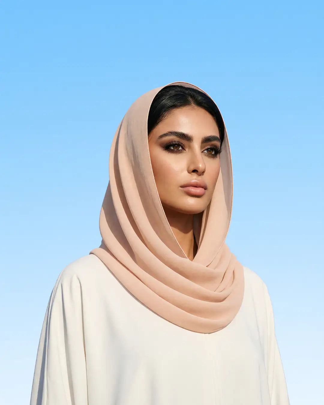 A woman wearing a soft blush-beige draped hijab and a cream-colored abaya, photographed against a clear light blue sky background.