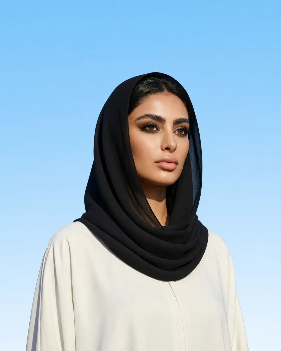 A woman wearing a black sheer and opaque double-layered hijab with a white abaya, set against a clear blue sky background.