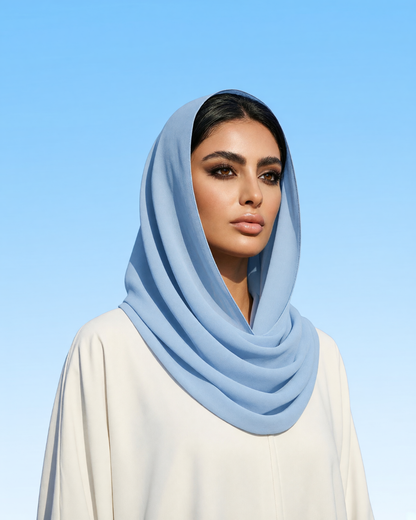 A woman wearing a light blue draped hijab and a cream-colored abaya against a clear blue sky background.