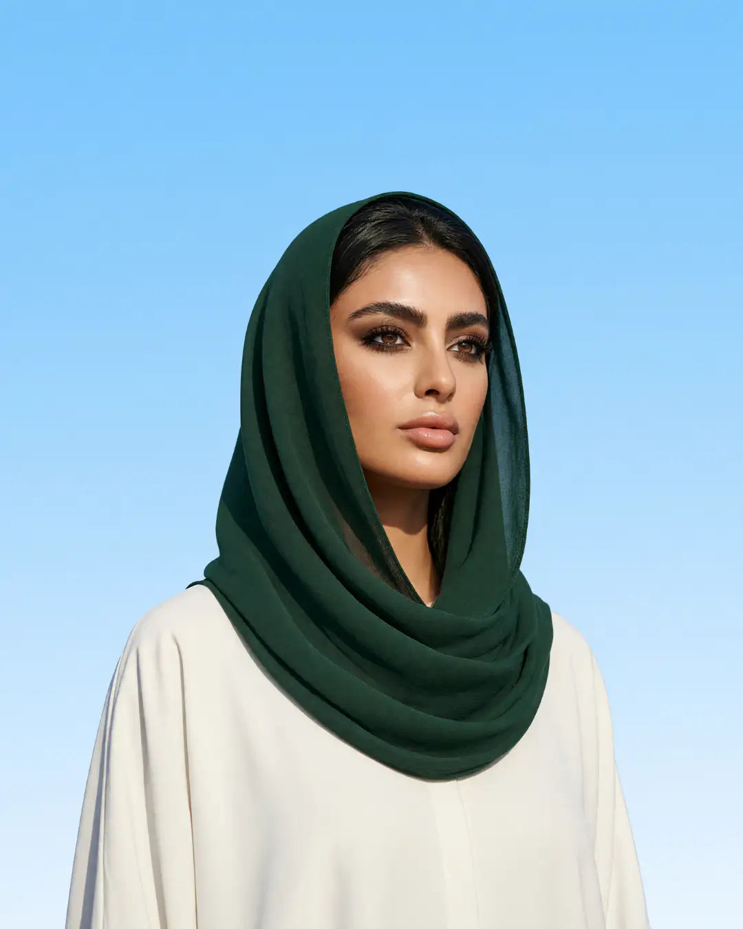 A woman wearing a dark forest green hijab and a white abaya, photographed against a clear light blue sky background.
