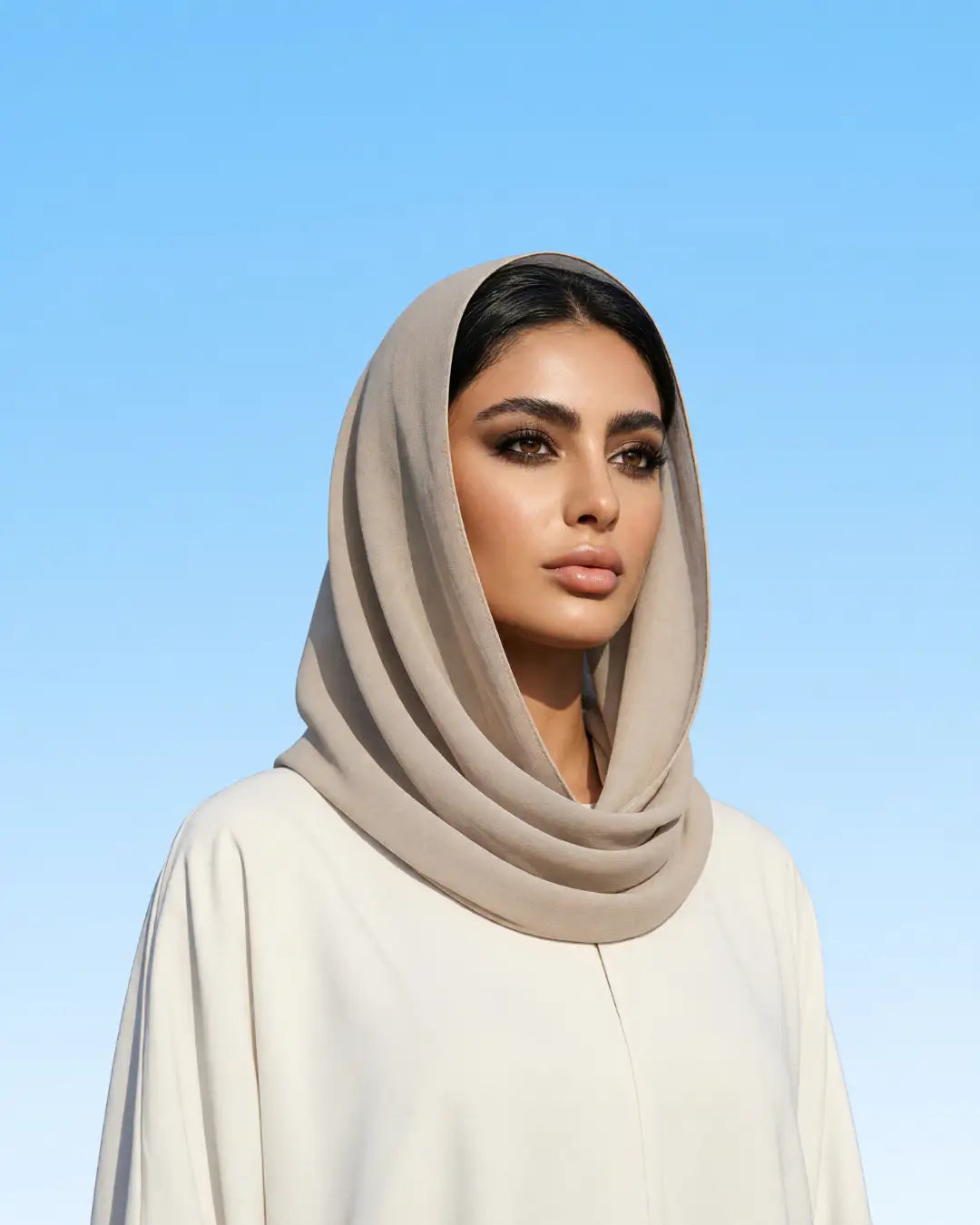 A woman wearing a draped taupe-beige hijab and a cream-colored abaya, photographed against a clear light blue sky background.