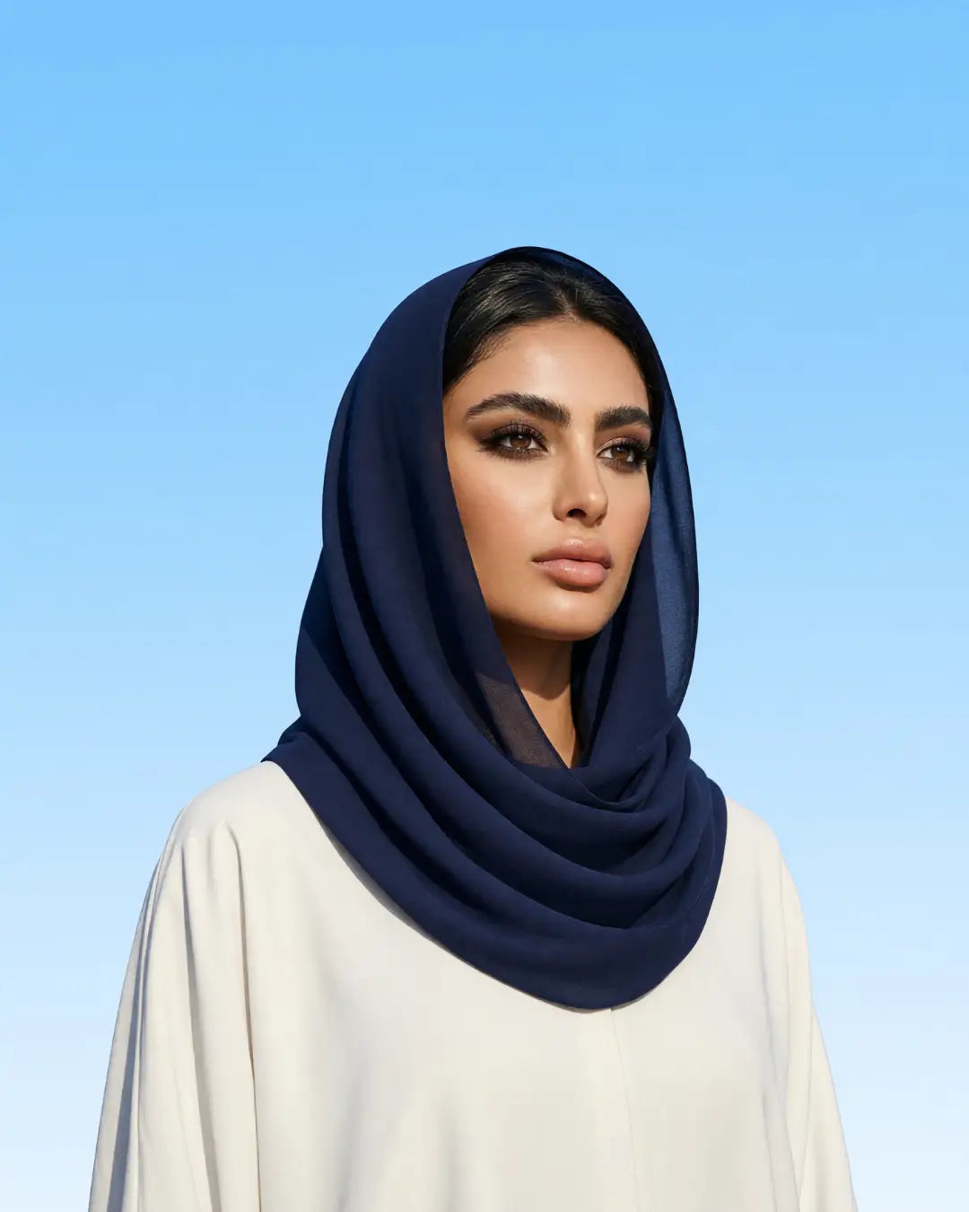 A woman wearing a dark navy blue draped hijab and a cream-colored tunic, set against a clear blue sky background.