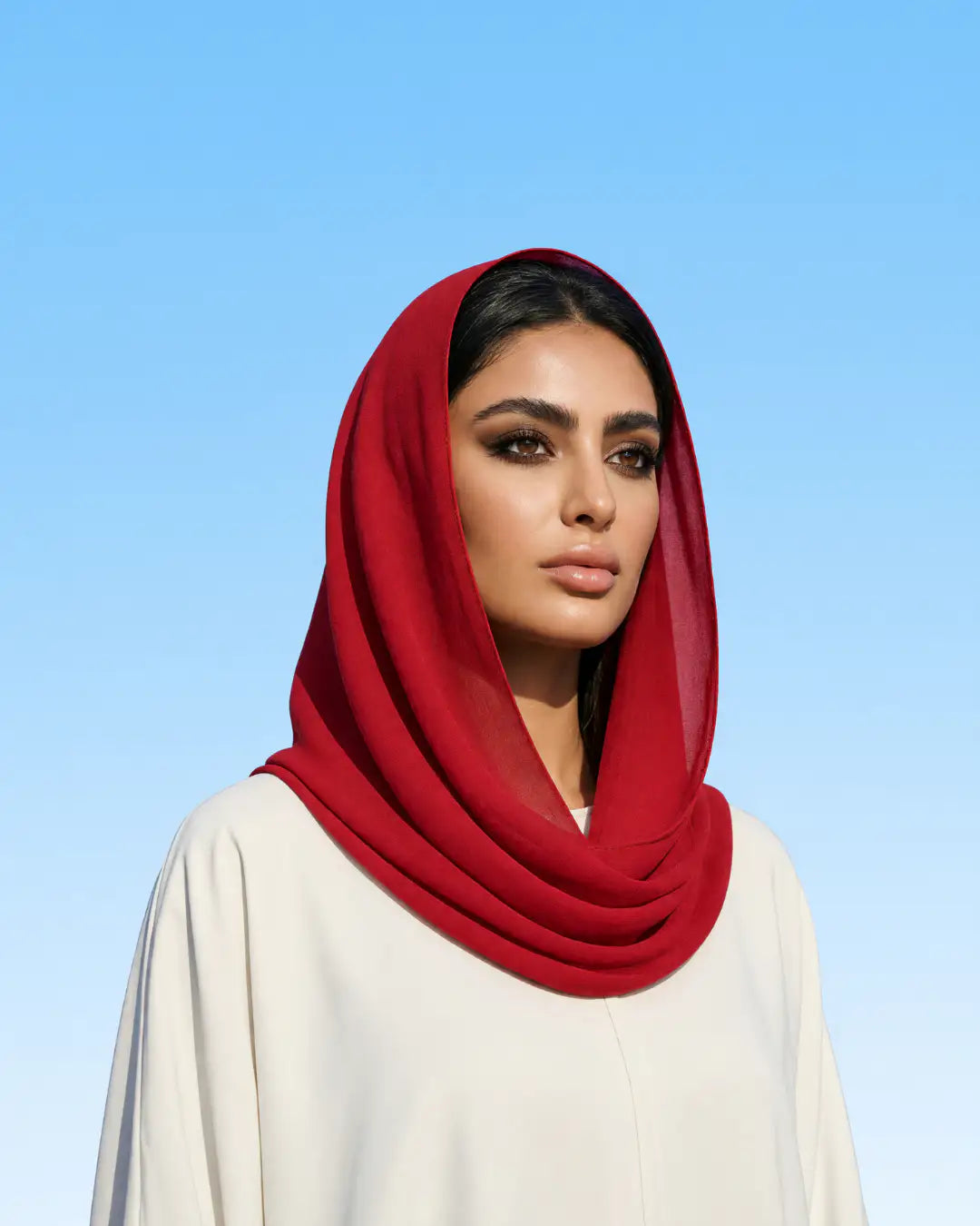 A woman wearing a deep red chiffon hijab and a cream-colored abaya, photographed against a clear blue sky of jeddah background.