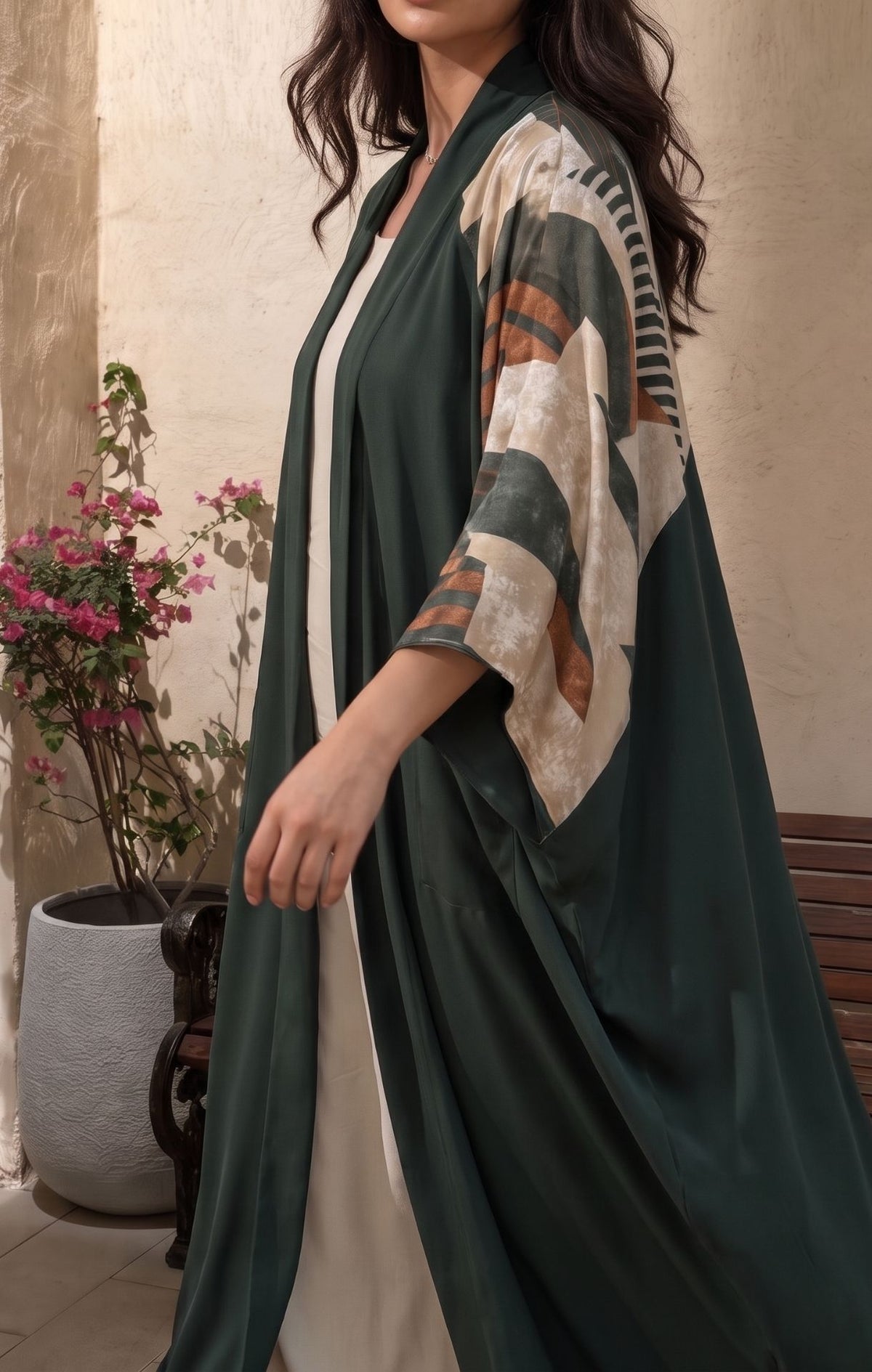 A woman wearing a dark green open-front kaftan-style robe with wide sleeves featuring a cream, brown, and green geometric pattern, posed in front of a textured beige wall with a potted plant.