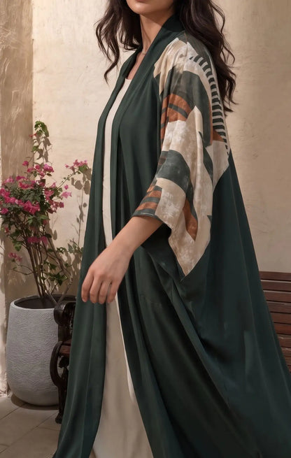 A woman wearing a dark green open-front kaftan-style robe with wide sleeves featuring a cream, brown, and green geometric pattern, posed in front of a textured beige wall with a potted plant.