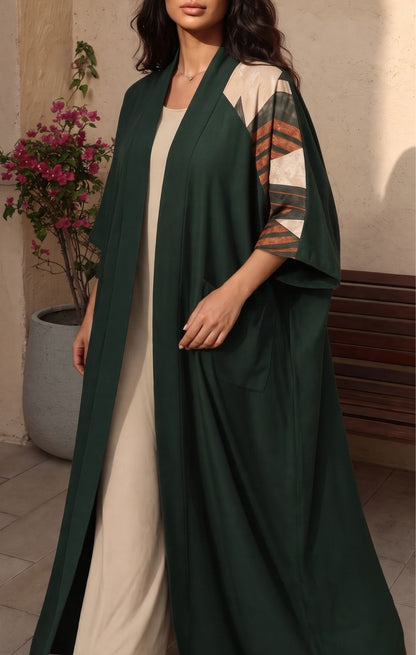 A woman wearing a long dark green open-front kaftan or abaya dress over a cream dress, with patterned three-quarter sleeves, standing in an outdoor patio setting.
